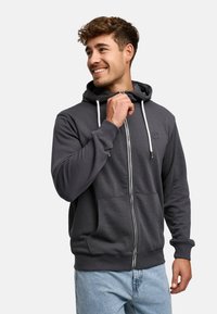 Grå zip-up hoodie i mjukt material med vita dragsnören, framfickor och ribbade muddar. Har en liten logotyp på bröstet.
