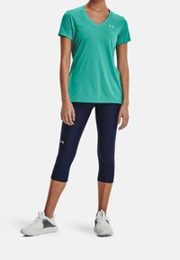 Lekki, turkusowy t-shirt sportowy z dekoltem w kształcie litery V, w połączeniu z granatowymi legginsami 7/8. Biały logo na zarówno na t-shircie, jak i legginsach. Model nosi białe trampki.