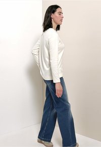 Witte longsleeve top in een ontspannen pasvorm, gecombineerd met een wijde spijkerbroek in blauw. De outfit heeft een eenvoudig ontwerp zonder patronen of accenten.