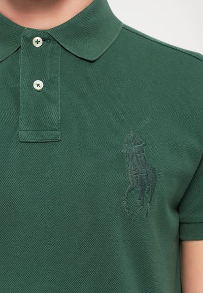 Polo de color verde oscuro con una tela texturizada, que presenta un cierre de dos botones y un logotipo bordado de un caballo y jinete en hilo a juego.