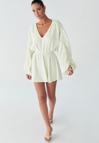 Een vrouw loopt zelfverzekerd in een lichte, geplooide witte jurk met een V-hals en lange mouwen, gecombineerd met witte slippers.