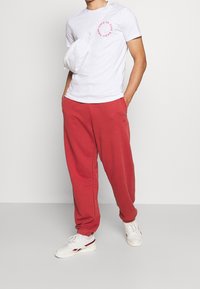 Vit T-shirt med rosa logga, kombinerad med röda sweatpants. Accessoarerna inkluderar en liten vit crossbody-väska och vita sneakers med röda detaljer.