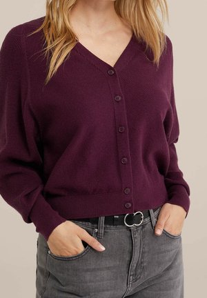 Cardigan a maglia di colore viola scuro, con scollatura a V e cinque bottoni, abbinato a jeans grigi. Presenta polsini a coste e una vestibilità comoda.