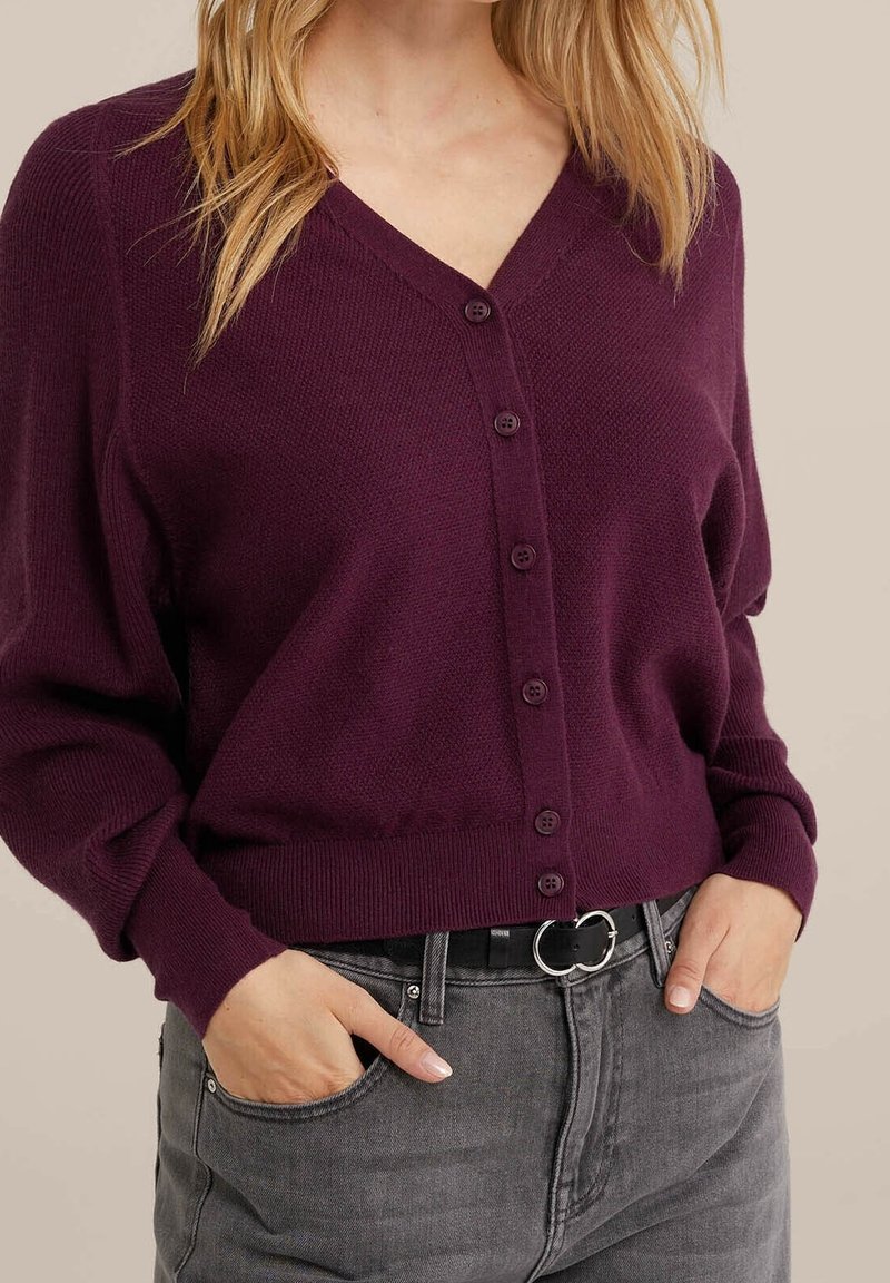 Cardigan en tricot violet foncé avec un col en V et cinq boutons, associé à un jean gris. Il présente des poignets côtelés et une coupe décontractée.