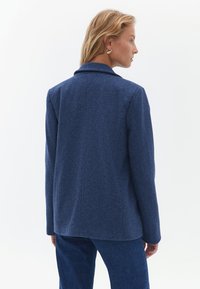 Blazer z vlněné směsi v tmavě modré barvě, s klasickým límcem, pohodlným střihem, strukturovanou tkaninou a rovnou zadní stranou s jemnými švy.