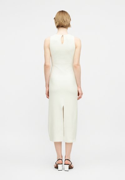 Γυναίκα με κοντά ξανθά μαλλιά που φοράει λευκό αμάνικο μεσαίο φόρεμα με σκίσιμο στο πίσω μέρος και λεπτομέρεια με άνοιγμα, καθώς και μαύρα σανδάλια με χοντρό τακούνι.