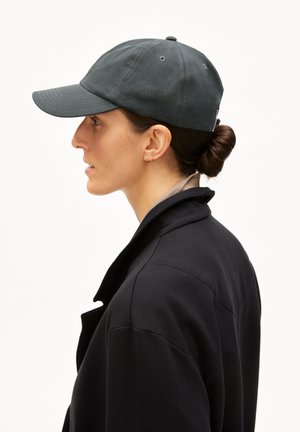 Femme portant une casquette de baseball sombre, de petites boucles d'oreilles dorées en forme d'anneau, et un manteau noir, les cheveux attachés en chignon, vue de profil sur un fond blanc.