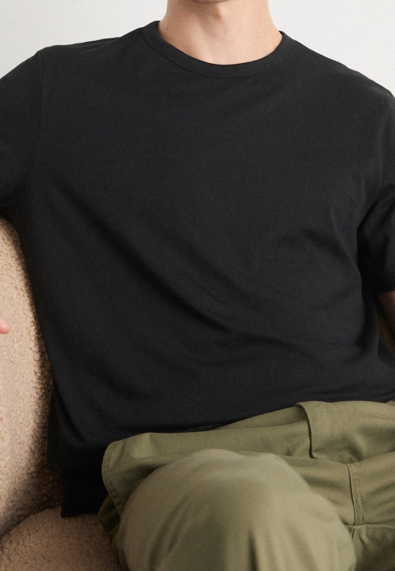 Mustat puuvillaiset t-paita lyhyillä hihoilla, joissa on pyöreä kaulus, yhdistettynä oliivinvihreisiin housuihin. Kangas on sileä ja tasainen.