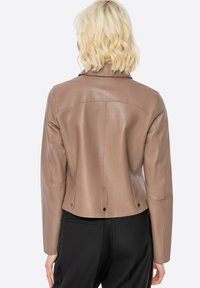 Blonde persoon in een strakke bruine leren jas en zwarte broek, gezien van achteren tegen een witte achtergrond.