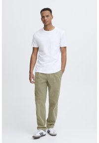 T-shirt en coton blanc, pantalon cargo vert clair, baskets blanches et grises avec des bandes noires. Le mannequin se tient contre un fond clair uni.