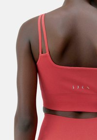 Reggiseno sportivo rosso con spalline sottili, dettagli testurizzati e una finitura liscia. Presenta un logo discreto sul retro, abbinato a un pantalone a vita alta coordinato.