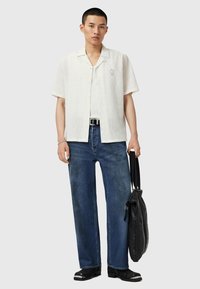 Chemise blanche à manches courtes, tissu texturé, logo subtil ; jeans bleu à coupe ample ; sac noir en bandoulière ; chaussures noires avec accents argentés.