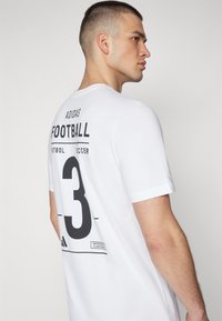T-shirt bianco in cotone a maniche corte, con una stampa grafica nera che reca la scritta "ADIDAS FOOTBALL" e il numero "3" sul retro.
