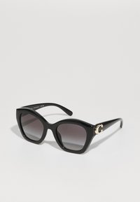 Lunettes de soleil noires avec une forme audacieuse en œil de chat, dotées de verres gris dégradés et d'un accent doré en "G" sur le côté. Fabriquées en plastique brillant.