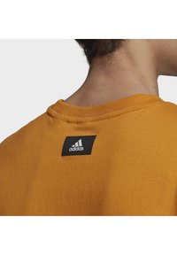 Orange pullover-tröja med ribbad rund hals; har en svart fyrkantig etikett med vit Adidas-logga på baksidan nära nacken.