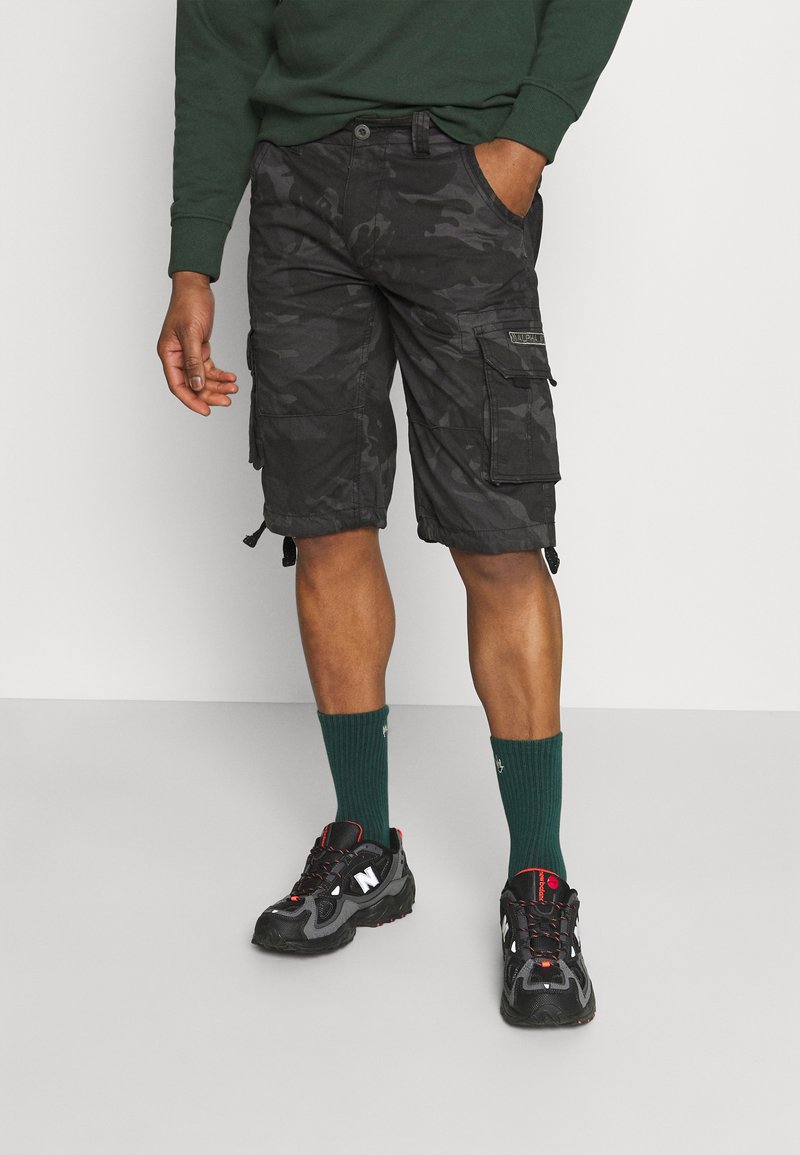Pantaloni cargo camo neri in tessuto con molteplici tasche e cordini regolabili all'orlo, abbinati a calzini verdi e sneakers nere.