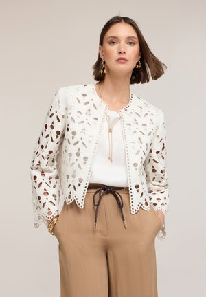 Kobieta ubrana w białą kurtkę z ażurowej koronki na białym topie, beżowe spodnie z wysokim stanem i czarnym sznurkiem, złote kolczyki, naszyjnik i bransoletki.