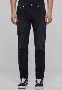 Jeans de mezclilla negra con un corte ajustado, que cuentan con cinco bolsillos, un ligero desgaste y un cierre de botón. Combinados con zapatillas negras y blancas.