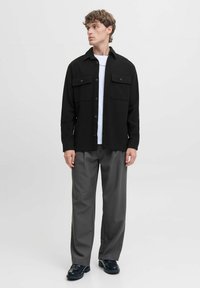 Camicia nera con bottoni e due tasche sul petto, abbinata a pantaloni grigi. Il modello sta in piedi con le mani ai lati, indossando scarpe nere.