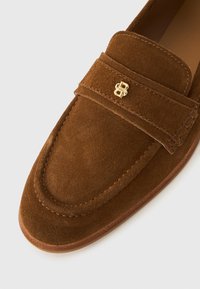 Brown suede loafers with a rounded toe and stitched accents, featuring a gold logo on the strap. Smooth texture and minimalistic design.