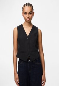 Junge Frau mit geflochtenem Haar trägt eine ärmellose schwarze Knopfjacke und dunkelblaue Jeans und steht vor einem schlichten weißen Hintergrund.