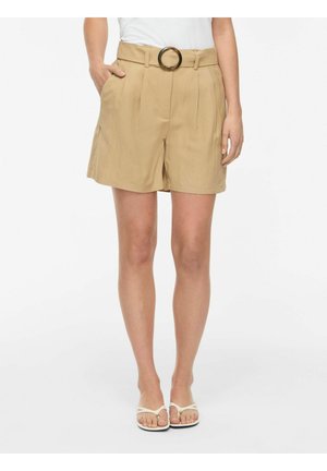 Femme portant un short beige taille haute avec une grande boucle de ceinture ronde, un haut blanc et des sandales blanches, debout avec une main dans la poche.
