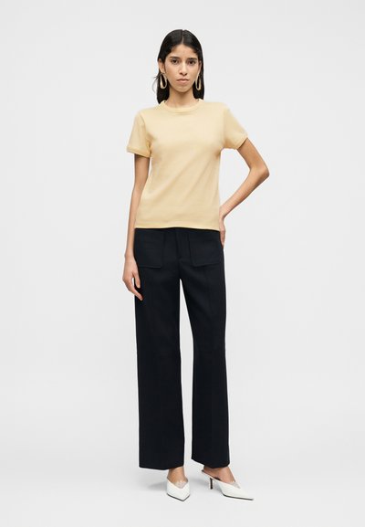 Βαμβακερό t-shirt με κοντά μανίκια σε ανοιχτό κίτρινο, συνδυασμένο με μαύρο ψηλόμεσο παντελόνι που διαθέτει δύο μπροστινές τσέπες και λευκά ψηλοτάκουνα mules.