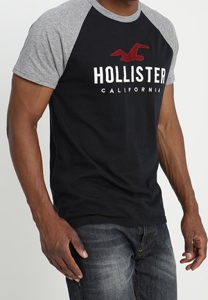 Mand iført sort og grå Hollister California kortærmet skjorte og falmede blå jeans, stående mod en enkel lys baggrund.