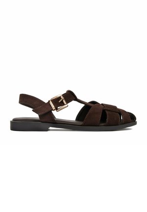Sandalia plana de ante marrón con punta cerrada, laterales abiertos, correa ajustable con hebilla dorada y suela baja negra.