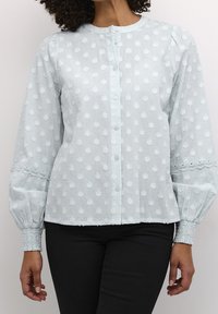 Hellblaue Bluse mit strukturiertem Muster, Knopfleiste vorne, Biesen-Details und Spitzeneinsätzen an den Ärmeln. Weicher Stoff mit Puffärmeln.