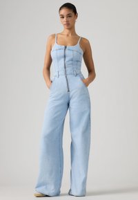 Γυναίκα στέκεται με τα χέρια στις τσέπες φορώντας ολόσωμη φόρμα από ανοιχτό μπλε τζιν με φαρδιά μπατζάκια, εμπρός φερμουάρ και λεπτές τιράντες, σε απλό φόντο.