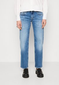 Svetle modré rovné džíny s bledými oblastmi a jemným opotřebením. Mají klasický pětivočkový design a zipový klopení. Nosené s černými botami.