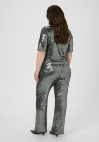 Glittrig jumpsuit i silver med korta ärmar och avslappnad passform. Urringade byxor och slät textur som reflekterar ljus. Svarta klackskor på. 