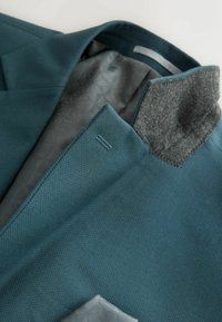 Turkusowa marynarka z teksturowanej tkaniny i szarym, filcowym kołnierzem, z wzorzystą szaro-skalną podszewką i widocznymi detalami szwu.