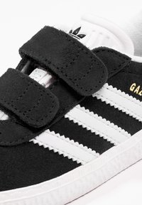 Svarta mocka sneakers med vita ränder, kardborreband och texturerad gummisula. Har en vit logotyp på tungan och guld detaljer.