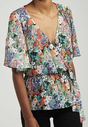 Femme portant un chemisier portefeuille coloré à motifs floraux avec des manches volantées et une taille nouée, associé à un pantalon noir.