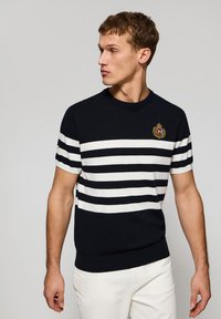 Marineblauer, gestrickter Kurzarmpullover mit weißen horizontalen Streifen, rundem Halsausschnitt und gesticktem Logo auf der Brust. Glatte Textur.