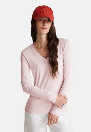 Chemise à manches longues de couleur rose clair avec un décolleté en V, fabriquée en tissu doux, associée à une casquette rouge arborant un design de logo en relief. Le modèle pose avec une bague.