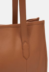 Sac fourre-tout en cuir marron avec une surface texturée, deux poignées robustes et des coutures visibles le long des bords. Design sans fermeture, matériel minimal.