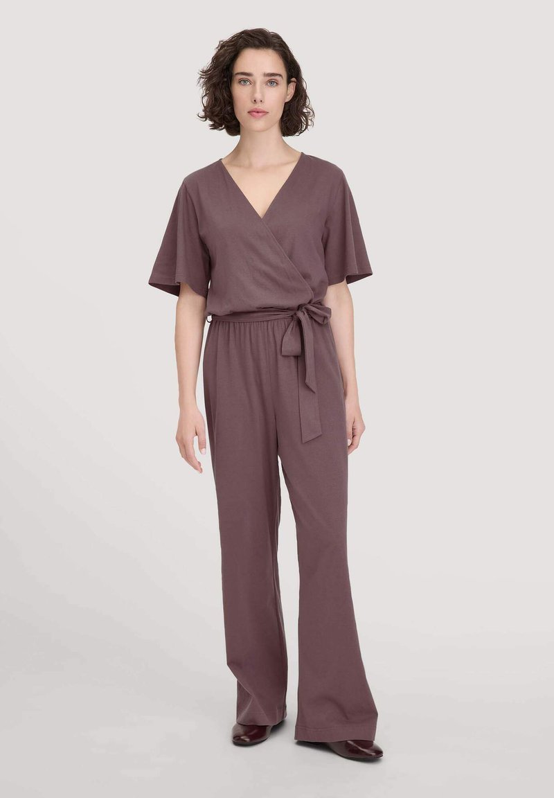 Vrouw met kort krullend haar, gekleed in een mauve wikkel-jumpsuit met brede pijpen en een strikceintuur, staande tegen een lichte achtergrond.