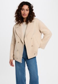 Mujer con cabello rizado lleva un abrigo beige de doble botonadura, un suéter claro y jeans azules, posando contra un fondo blanco liso.