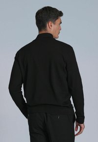 Pull en maille noir avec un col montant et des poignets côtelés. Le dos présente une texture lisse, avec une silhouette classique et ajustée.