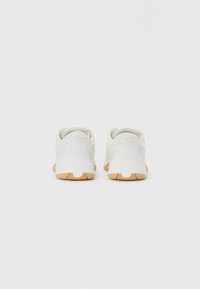 Et par hvide sneakers med brune såler, set fra bagsiden på en ensfarvet lys grå baggrund.