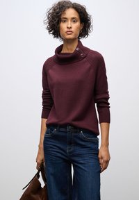Burgunderfarbener Strickpullover mit hohem Kragen und Knopfdetails; kombiniert mit blauen Jeans. Das Model hält eine braune Handtasche.