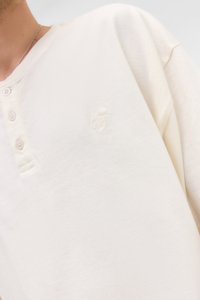 Cream-colored henley shirt made of soft fabric with three buttons and a textured embroidered logo on the left chest. Short sleeves.