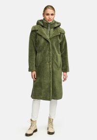 Groene met bont gevoerde jas met een geknipt kraag, twee knopen en voorkanten, gelaagd over een groene hoodie met ritssluiting. Witte broek en beige laarzen.