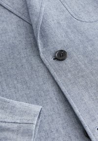 Gros plan sur un blazer texturé bleu clair avec un bouton foncé fermé près du revers et une couture de poche visible.