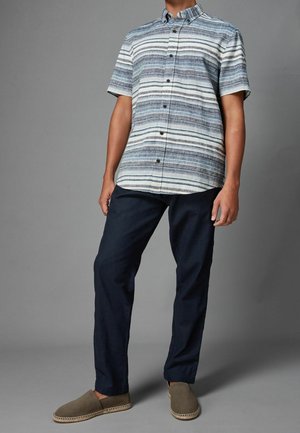 Camicia a maniche corte con bottoni, a strisce orizzontali blu e bianche, abbinata a pantaloni scuri blu navy e scarpe slip-on grigie.