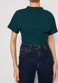 Tyrkysový přiléhavý top s vysokým límcem a krátkými rukávy, doplněný temně modrými džínami s vysokým pasem. Tkanina vypadá hladce a pružně.