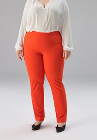 Rote, eng anliegende Hose aus glattem Stoff, kombiniert mit einer weißen Bluse mit V-Ausschnitt und elastischen Bündchen. Schwarze Ballerinas vervollständigen das Outfit.